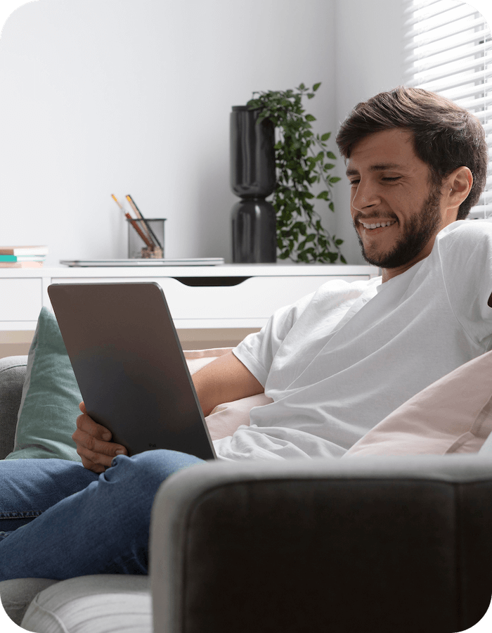 A young man in a white shirt sitting on a sofa, opening his laptop which is connected to Eweka’s VPN for advanced threat protection settings.