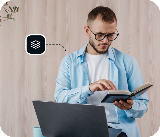 A man wearing glasses is referencing a book in front of an open laptop to symbolize the ease with which Usenet users are able to locate Usenet articles from the Eweka Usenet archive.