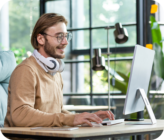 A man wearing glasses has a pair of headphones around his neck and is typing on a desktop computer, accessing the large Usenet archive that Eweka has to offer thanks to its massive article retention rates.