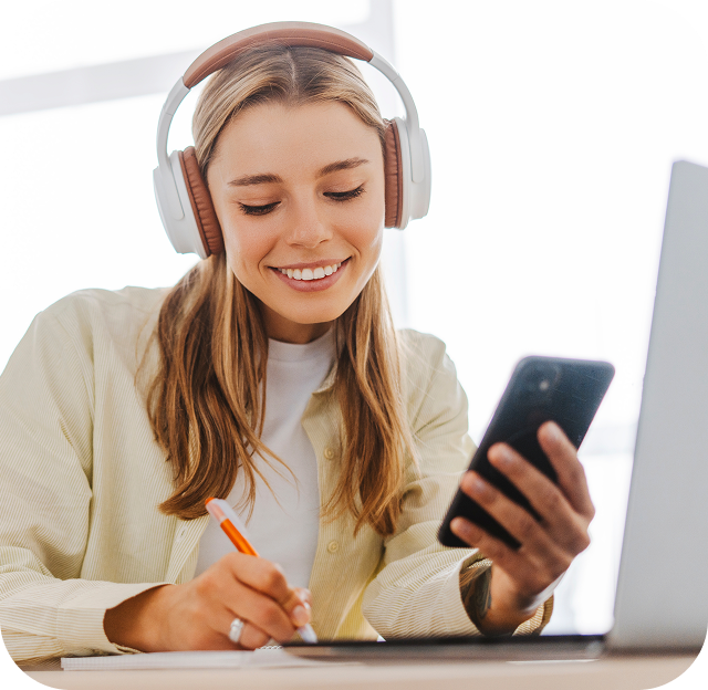 A young woman wearing headphones and holding her cellphone is sitting in front of an open laptop using Eweka Internet Services and taking notes in a notepad.