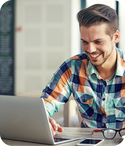 A man in a plaid shirt is smiling at an open laptop beside a smartphone, his glasses sitting nearby.