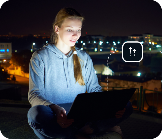 A young woman sits outside, at night, on a laptop, showing that users can connect to Eweka day and night and enjoy the most uptime of any Usenet provider. 