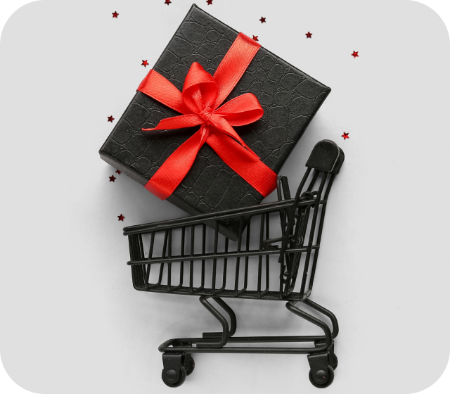 A black shopping cart in front of a white wall with a large package inside. The package is wrapped in black paper and has a red bow on it.