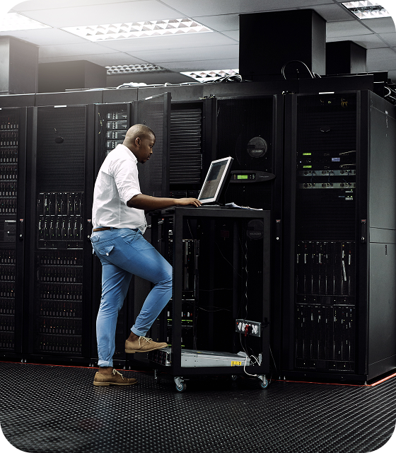 A man in a white shirt stands in a large Usenet server room, working on a laptop to ensure maximum efficiency for Usenet users.