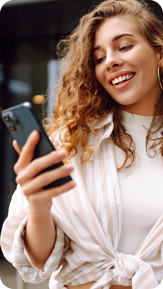A smiling woman in a white shirt with a cream and white striped overshirt tied at the midriff is looking at her cellphone.
