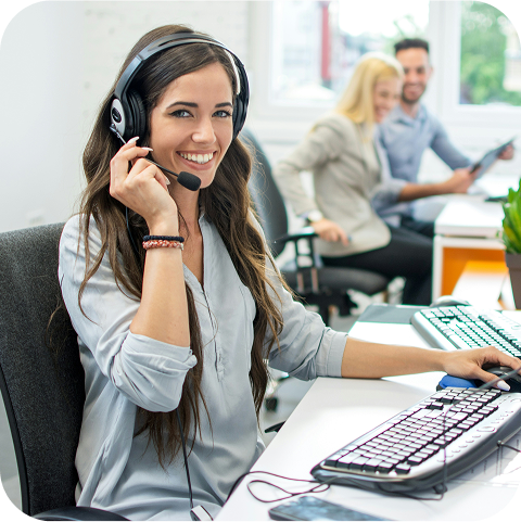 A woman wearing a headset is sitting in an open-concept office smiling at the camera.