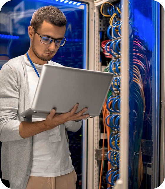 A man wearing a grey cardigan is holding a laptop on his right hand and typing with his left. He is standing in what appears to be a Usenet service provider data center.