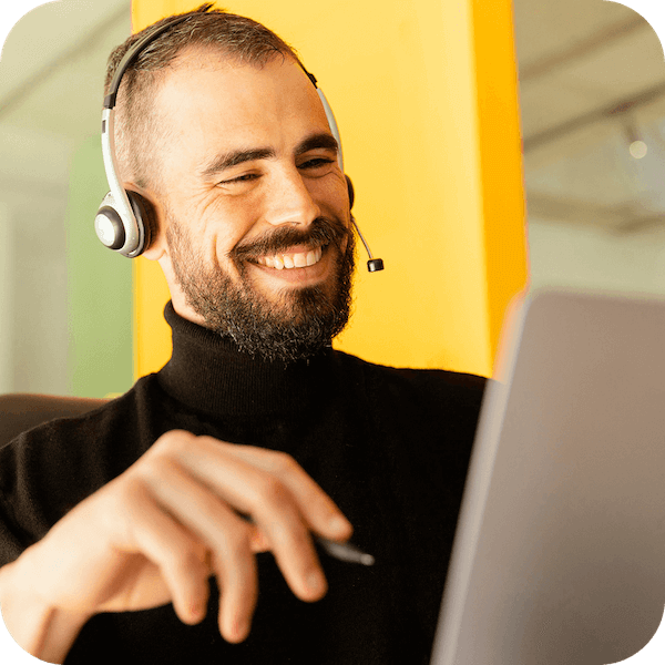 A male customer support agent in a black turtleneck is smiling and sitting in front of a laptop while wearing a headset to speak with a customer.