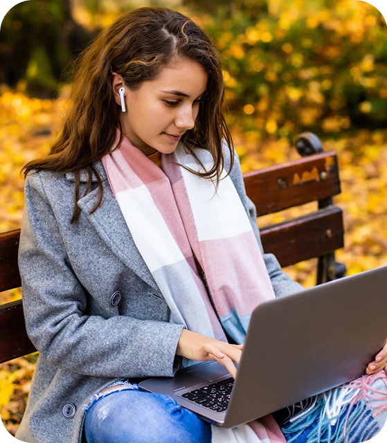 Een vrouw met een grijs-witte-roze sjaal, een grijze mantel en gescheurde jeans zit op een parkbank met een laptop op schoot en AirPods in haar oren.