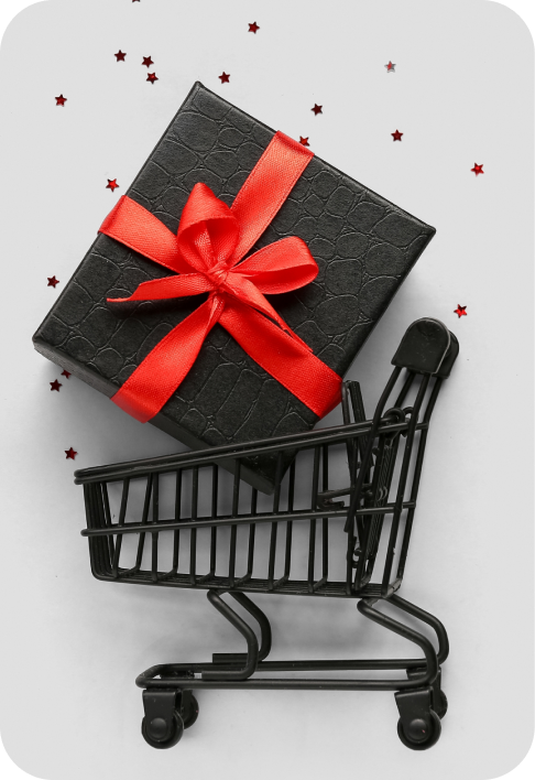 A black shopping cart in front of a white wall with a large package inside. The package is wrapped in black paper and has a red bow on it.
