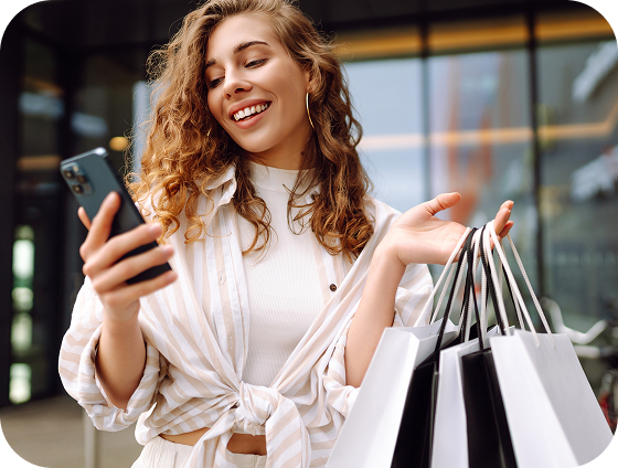 A smiling woman in a white shirt with a cream and white striped overshirt tied at the midriff is looking at her cellphone.