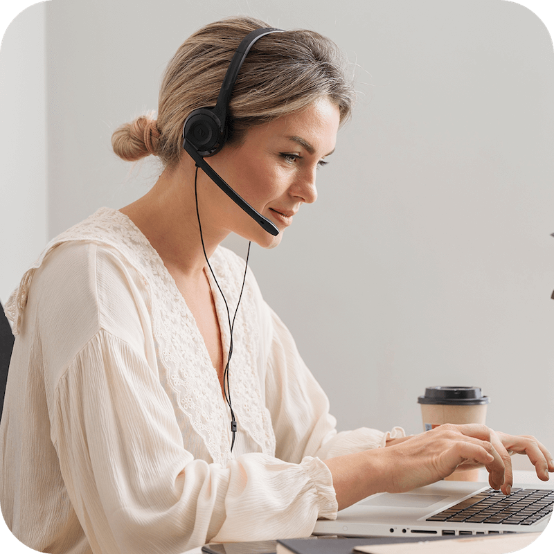 Een vrouw in een wit shirt helpt een klant via een headset terwijl ze op een laptop typt.