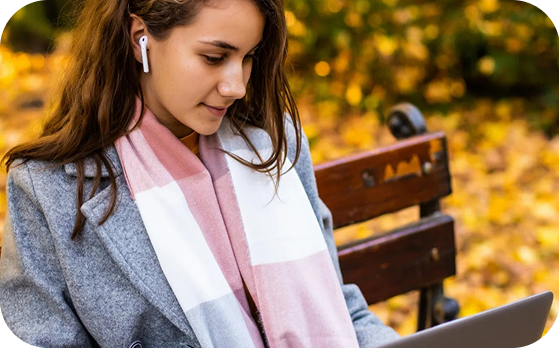 A woman wearing a grey, white, and pink scarf, a grey pea coat, and ripped jeans is sitting on a park bench with a laptop on her lap and Airpods in her ears.